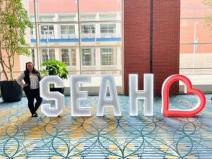 A woman standing next to a sign that says SEAH with a heart at the end.